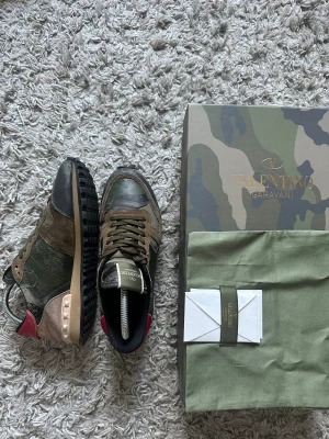 Valentino Rockrunner sneakers camo - Säljer ett par Valentino Rockrunner sneakers i camo-mönster med gröna, bruna och svarta toner. Skorna har detaljer i mocka och skinn, coola nitar vid hälen och röd accent baktill. Klassisk snörning och platt sula. Kommer ej med originalkartong och dustbag. Kan dock skicka med vid förhandling 