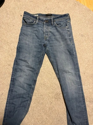 Blå jeans från Jack & Jones - Snygga blå jeans från Jack & Jones med klassisk femficksdesign och diskret tvättad look. Modellen har raka ben och normal passform, med detaljer som knappgylf och läderpatch bak i midjan. Perfekta för dig som gillar stilrena och bekväma jeans.