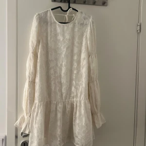 Vit kortklänning med volangärm - Superfin helt oanvänd vit kortklänning från Gina Tricot med blommigt mönster i tyget. Klänningen har långa volangärmar, rund halsringning fram och djupare rygg med knytband. Lätt och flowy känsla, perfekt för sommarens alla tillfällen.
