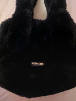 Svart pälsväska från Love Moschino - Supermjuk och fluffig svart axelväska från Love Moschino i fuskpäls. Väskan har breda handtag och en stilren guldlogga framtill. Perfekt för dig som vill ha en statement-väska med lyxig känsla och trendig look. Nypris: 2000