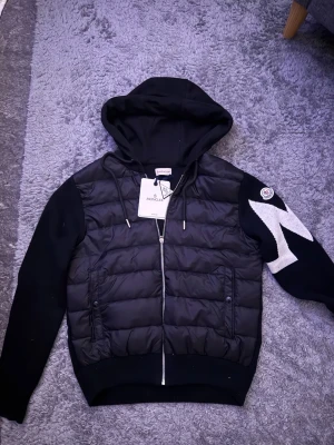 Moncler Cardigan - Svart pufferjacka från Moncler med huva och dragkedja. Jackan har quiltad front i dun och ärmar i stickad bomull med vit zigzag-detalj och Moncler-logga på ena ärmen. Snygg och sportig look, perfekt för kyliga dagar. Mer bilder skickas privat! Tveka inte på att fråga om du undrar något!