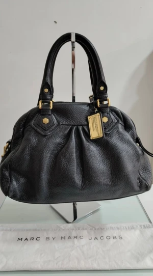 Svart handväska från Marc by Marc Jacobs - Cool svart handväska i mjukt skinn från Marc by Marc Jacobs med guldfärgade metalldetaljer och dekorativ bricka framtill. Väskan har dubbla handtag, dragkedja upptill och ett unikt innerfoder med grafiskt mönster i svart och vitt. Köpt för 4000kr som nyskick 