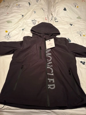 Svart vindjacka från Moncler med huva - Svart vindjacka från Moncler med stor logga längs dragkedjan. Jackan har huva med dragsko, flera dragkedjeförsedda fickor och en clean, sportig look. Materialet är lätt och syntetiskt, perfekt för blåsiga dagar. Snygg och modern passform.