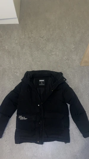 Svart Dunjacka från Hoodrich storlek L (Junior) - Säljer en svart Hoodrich-jacka i storlek L (Junior). Jackan är vadderad och varm, perfekt för höst och vinter. Den har en hög krage och huva för extra skydd mot kyla.  Framtill finns flera praktiska fickor, varav en har texten “FROM NOTHING TO SOMETHING™”. Jackan stängs med dragkedja och tryckknappar. Färgen är matt svart och passformen är normal till något rymlig – typisk puffer-stil. Passar dig som vill ha en stilren, varm och street-inspirerad vinterjacka. 