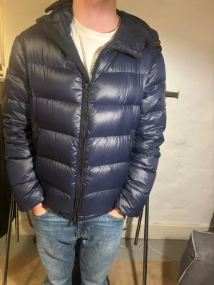 Mörk blå jacka från Peuterey - Snygg mörkblå dunjacka från Peuterey med huva och två sidofickor med dragkedja. Jackan har ett quiltat mönster och är tillverkad i glansig polyester. Perfekt för kalla dagar och har en stilren look med svarta detaljer och märkets logga på ärmen. Modellen på bilden är 179 cm lång, storlek M, mycket bra skick, nytt pris 5000kr mitt pris 1999kr✅. Hör av dig vid funderingar 📩