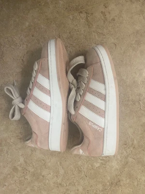 Adidas Campus rosa sneakers mocka - Säljer ett par Adidas Campus sneakers i ljusrosa mocka med vita klassiska ränder och vit sula. Skorna har snörning och rund tå, samt Adidas-logga på hälen och plösen. Perfekta för dig som gillar en clean och sportig look med en touch av pastell.