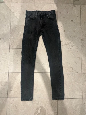 Nudie jeans slitningar 31/34 - Svarta jeans från Nudie med skinny passform och klassisk femficksdesign. Jeansen har slitningar och hål på ena benet för en edgy look. Bakfickorna har Nudies signaturbroderi i svart. Perfekt för dig som gillar streetstyle.