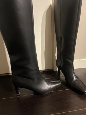 Svarta höga stövlar med klack - Snygga svarta stövlar i skinn med spetsig tå och smal klack. Stövlarna är knähöga och har en stilren, minimalistisk design som passar perfekt till både jeans och kjol. Materialet är slätt och ger en lyxig känsla.