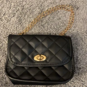 Svart quiltad axelväska med guldkedja - Snygg svart axelväska i quiltad design med guldfärgad kedja och vridlås. Väskan har en klassisk look och är tillverkad i skinnimitation. Perfekt storlek för det viktigaste och ger en lyxig känsla till din outfit.