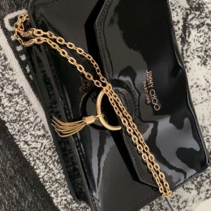 Svart lackad kuvertväska Jimmy Choo - Snygg svart kuvertväska från Jimmy Choo Parfums i glansigt lackmaterial. Väskan har guldfärgad kedja och dekorativ tofsdetalj framtill. Perfekt storlek för det viktigaste och stängs med magnetlås. Lyxig och trendig accessoar med exklusiv känsla. Priser går att diskutera🫶🏼🥰