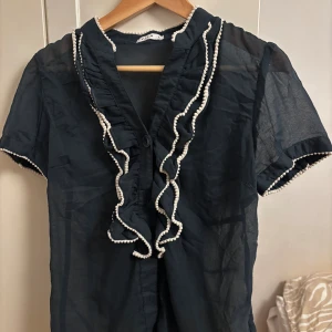 Marinblå blus med volang från Vila - Marinblå, tunn blus från Vila med korta ärmar och vita volangdetaljer längs krage och knappslå. Blusen har en lätt transparent känsla och dekorativa små bollar längs volanger och ärmslut. Perfekt för dig som gillar unika detaljer.