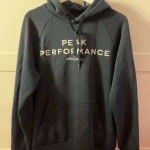 Mörkblå hoodie från Peak Performance - Snygg mörkblå hoodie från Peak Performance med vit logga på bröstet. Tröjan har huva med snörning och en stor magficka framtill. Perfekt för chill dagar och har en klassisk, loose passform. Materialet är mjuk bomull och polyester.