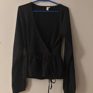 Svart omlottopp från Nelly - Svart omlottopp från Nelly med långa ärmar och knyt i midjan. Toppen har v-ringning och en lätt rynkad effekt vid midjan för en snygg siluett. Perfekt att styla med jeans eller kjol för en trendig look.