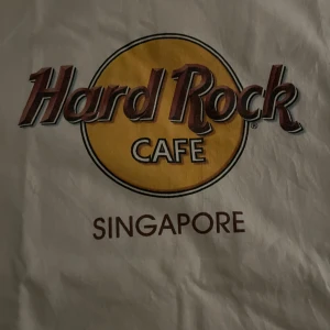 Hard Rock Cafe t-shirt - Vit t-shirt i bomull med klassiskt Hard Rock Cafe-tryck i gult och brunt framtill. Finns med olika städer som Singapore, Makati, Sosua och Ft. Lauderdale. Perfekt för dig som gillar ikoniska loggor och samlarplagg. Skriv till mig vilken ni skulle vara intresserad av innan ni köper💕olika storlekar på olika tröja🥰