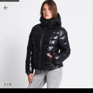 Svart glansig pufferjacka med huva lager 157 - Svart pufferjacka med glansig finish och stor, mysig huva. Jackan är väldigt fin och gar en bra passform! Håller värme bra nu till kallare tider. Har bara använt den ungefär två gånger.