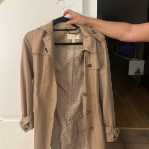 Beige trenchcoat från Michael Kors s - Snygg beige trenchcoat från Michael Kors med klassisk design, knappar framtill, axelklaffar och bälte i midjan. Fodret har MK-logga över hela insidan. Perfekt för dig som vill ha en stilren och tidlös jacka med coola detaljer.