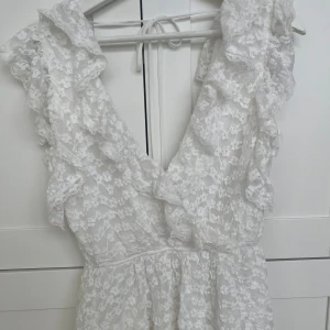 Vit spetsklänning från Nelly M - Superfin vit spetsklänning från Nelly med volangdetaljer och v-ringning. Klänningen har omlottkänsla upptill, knytband i nacken och är ärmlös. Perfekt för sommarens alla tillfällen när du vill känna dig extra söt.