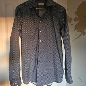 Mörkblå prickig skjorta från Eton - Snygg mörkblå skjorta från Eton med vita prickar och klassisk krage. Slim fit-modell med långa ärmar och vita knappar. Tillverkad i bomull för en skön känsla och stilren look. Fattas en knapp framtill.