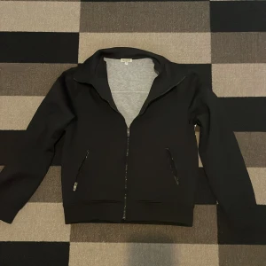 Svart ziptröja från Kenzo med logga - Svart långärmad ziptröja från Kenzo med vit panel och stor Kenzo Paris-logga på ryggen. Tröjan har dragkedja framtill, två fickor med dragkedja och en klassisk krage. Perfekt för dig som gillar streetstyle och vill sticka ut med en snygg logga.