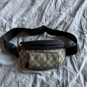 beige och svart axelväska/ magväska - Snygg magväska i beige canvas med klassiskt GG-mönster och svarta skinn- och textildetaljer. Väskan har justerbart band, dragkedjor och flera fack för smidig förvaring. Perfekt accessoar för en trendig streetstyle-look.