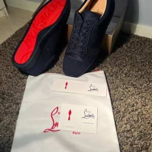 Mörkblå sneakers från Christian Louboutin - Snygga mörkblå sneakers från Christian Louboutin i mocka med klassisk röd sula och broderad logga på hälen. Skorna har rund tå, platt sula och snörning. Perfekta för dig som vill sticka ut med exklusiva detaljer och lyxig känsla.