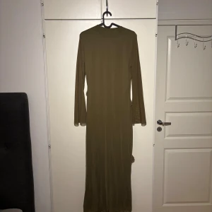 Olivgrön långklänning med volangdetaljer - Säljer en olivgrön långklänning med långa ärmar och rund halsringning. Klänningen har flera diagonala volangdetaljer framtill som ger en cool och trendig look. Materialet är tunt och faller snyggt längs kroppen. Perfekt för dig som gillar unika detaljer och stilren design. PRIS KAN ALLTID DISKUTERAS!!
