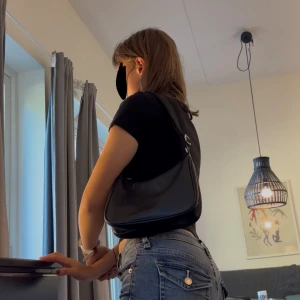 Svart axelväska i skinnlook - Snygg svart axelväska med stilren design och justerbart axelband. Väskan har en mjuk, slät yta i skinnimitation och en liten blomdetalj framtill. Perfekt storlek för mobil, plånbok och andra essentials. Har en fläck på ena sidan som inte stör något vid användning.