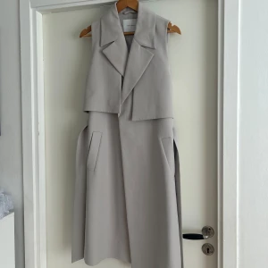 Beige lång väst från Ivy & Oak - Stilren beige lång väst från Ivy & Oak med breda slag, fickor och bälte i midjan. Västen har en snygg omlottkänsla och är perfekt för lager-på-lager. Materialet känns mjukt och har en lyxig look.