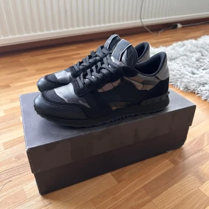 Valentino Garavani Rockrunner sneakers - Snygga Valentino Garavani Rockrunner sneakers i svart och grått med camouflagemönster. Skorna har detaljer i mocka och skinn, samt klassisk snörning och platt sula. Perfekta för dig som vill ha en exklusiv och edgy look med streetkänsla. Skick 7/10. Storlek 41. Box ingår