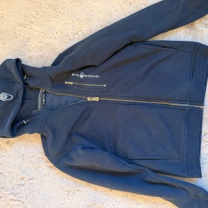 Mörkblå Sail Racing hoodiejacka - Mörkblå hoodiejacka från Sail Racing med dragkedja framtill och bröstficka med dragkedja. Jackan har huva med dragsko och mjukt fleecefoder på insidan. Perfekt för kyliga dagar och snygg logga på bröstet.