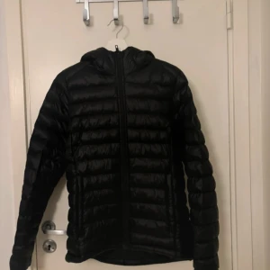 Svart dunjacka med huva - Säljer en svart dunjacka med huva och dragkedja framtill. Jackan har en quiltad design med horisontella sömmar och är lätt vadderad, perfekt för kyliga dagar. Passformen är normal och jackan har långa ärmar.