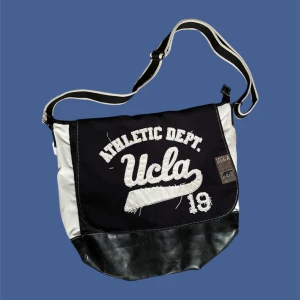 UCLA axelväska i svart och vitt - Cool axelväska från UCLA med broderad text och patch framtill. Väskan är svart med vita detaljer och har justerbar axelrem. Botten är i svart skinnimitation och resten i kraftig bomull. Perfekt för dig som gillar sportig och avslappnad stil.