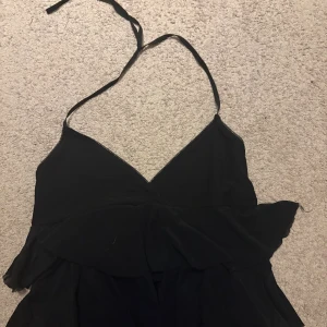 Svart halterneck  - Supersöt svart halterneck med smala axelband som knyts i nacken och v-ringad omlott framtill. Tröjan har flera lager volanger som ger en flowy och lekfull känsla. Perfekt för dig som gillar en feminin och trendig stil.