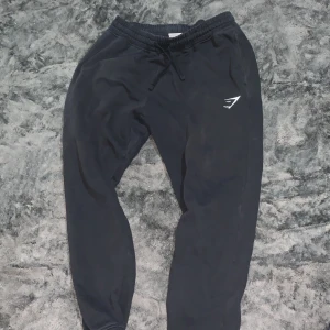 Svarta mjukisbyxor från Gymshark M - Svarta mjukisbyxor från Gymshark med elastisk midja och snörning. Byxorna har en vit Gymshark-logga på låret, ribbade muddar vid bensluten och fickor både fram och bak. Perfekta för chill eller träning.
