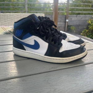 Nike Air Jordan 1 Mid svart/blå/vit - Säljer ett par Nike Air Jordan 1 Mid i svart, blått och vitt. Klassisk siluett med snörning, rund tå och vit sula. Skorna har blåa detaljer och swoosh, samt Air Jordan-logga på sidan. Perfekta för dig som gillar streetwear och sneakers med ikonisk design.