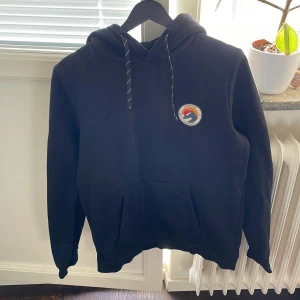 Svart hoodie från Everest med tryck - Svart hoodie från Everest med broderad logga på bröstet i rött, gult och vitt. Klassisk känguruficka framtill, snörning i huvan och långa ärmar. Mjuk och skön bomullsblandning, perfekt för chill dagar eller när du vill vara extra bekväm.