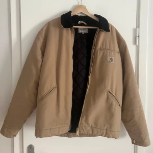 Beige Carhartt Detroit Jacka L - Säljer en beige Carhartt Detroit jacka med svart krage och quiltat foder. Jackan har dragkedja framtill, två snedställda fickor och klassisk Carhartt-logga på bröstet. Perfekt för höst och vinter, riktigt snygg och tidlös modell.