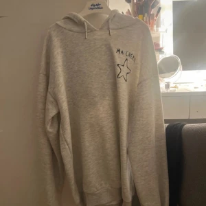 Grå hoodie från Gina Tricot med tryck - Mysig ljusgrå hoodie från Gina Tricot med svart text 'MA CHÉRIE' och en stjärna tryckt både fram och bak. Tröjan har huva, snörning och långa ärmar. Använd max 2 gånger, som ny. Storlek 158/164 men passar S