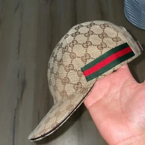 Beige Gucci keps med GG-mönster - Snygg beige keps från Gucci med klassiskt GG-monogram över hela kepsen och en ikonisk grön-röd rand på sidan. Justerbar rem i brunt skinn baktill. Materialet är bomull och skinn, perfekt för dig som vill ha en lyxig och trendig accessoar.