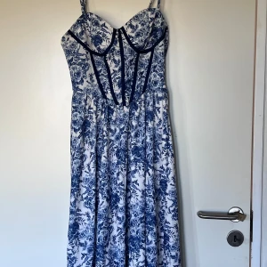 Blommig långklänning från SHEIN MOD - Supersnygg långklänning från SHEIN MOD med blått och vitt blommigt mönster. Klänningen har markerad byst med smala axelband och en flowy kjol. Materialet är mjukt och lätt polyester, perfekt för varma dagar. Den har en feminin siluett och retrovibbar.