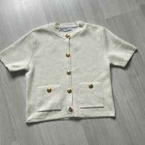 Vit kofta med guldknappar från Zara - Kortärmad vit kofta från Zara med snygga guldknappar framtill och två fickor. Koftan har en rak passform och är gjord i ett mjukt material som känns lyxigt. Perfekt för att lyfta en enkel outfit med lite extra detaljer.