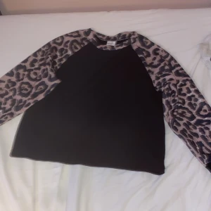 Svart tröja med leopardärmar - Cool svart tröja med långa ärmar i leopardmönster. Ärmarna och halsringningen har ett beige och svart leopardprint som ger en trendig look. Tröjan är gjord i mjuk polyesterblandning med lite stretch för extra komfort.