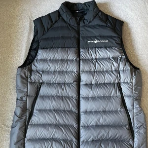 Svart och grå dunväst från Sail Racing - Snygg dunväst från Sail Racing i svart och grått med quiltad design. Västen har hög krage, dragkedja framtill och två fickor med dragkedja. Perfekt för lager-på-lager och har logga på bröstet samt en detalj på ryggen.