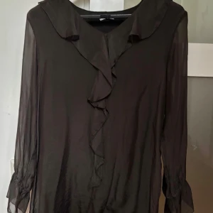 Brun blus med volang från Alberta Ferretti - Brun transparent blus från Alberta Ferretti med långa ärmar och snygg volang längs v-ringningen. Tillverkad i en lyxig mix av viskos och silke, vilket ger ett elegant och lätt uttryck. Perfekt för dig som gillar stilrena och feminina detaljer.