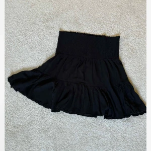 Svart volangkjol från 157, stl L - Svart kortkjol från 157 i storlek L. Kjolen har bred smock i midjan och flera volanger som ger en snygg och flowy look. Perfekt att styla med en croppad topp eller stickad tröja. Modellen heter Frannie.