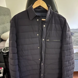 Blå quiltad pufferjacka med dragkedja - Stilren blå pufferjacka med quiltad design och raka linjer. Jackan har en klassisk krage, bröstficka med dragkedja och tryckknappar framtill. Perfekt för kyligare dagar och enkel att matcha med olika outfits.