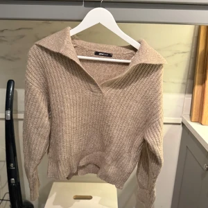 Beige stickad tröja från Gina Tricot - Säljer en beige stickad tröja från Gina Tricot med krage och v-ringning. Tröjan har lång ärm och en avslappnad passform, perfekt för lager-på-lager. Ribbstickade muddar vid ärmslut och nederkant ger en snygg detalj.