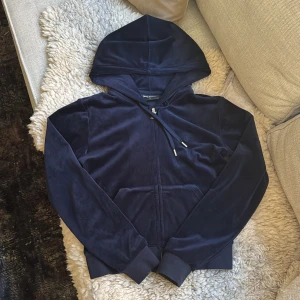Mörkblå velour hoodie Juicy Couture - Mörkblå hoodie från Juicy Couture i mjuk velour med huva, snörning och dragkedja framtill. Klassisk känguruficka och broderad JC-logga på bröstet. Perfekt för en chill och trendig look. Säljer även ett par byxor till!