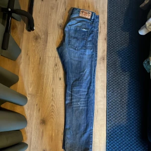 Diesel jeans - Säljer ett par blå Diesel Braddom jeans med snyggt slitna detaljer och klassisk femficksdesign. Jeansen har regular slim-carrot passform och coola slitningar framtill. 