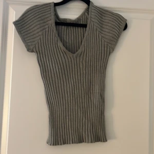 Grå ribbad topp med v-ringning - Säljer en grå ribbad topp med korta ärmar och v-ringning framtill. Toppen har en figurnära passform och är tillverkad i ett stretchigt material som sitter snyggt på kroppen. Perfekt att styla med jeans eller kjol för en clean look.